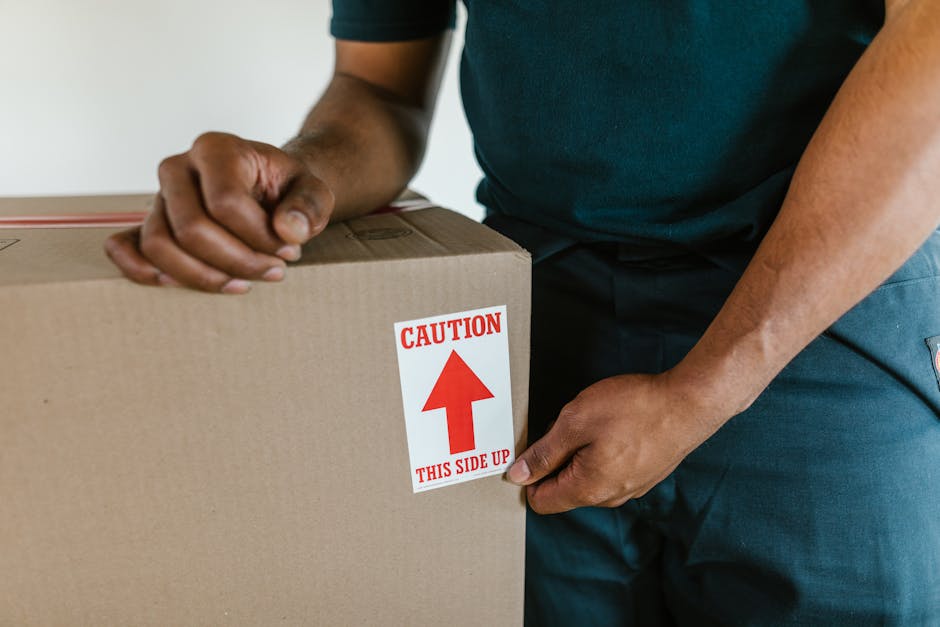 A person wearing a blue shirt and dark pants is holding a large cardboard moving box that has a red and white caution sticker on the side, indicating 'THIS SIDE UP' with an upward-pointing arrow, during a home relocation process. The individual is gripping the box with both hands, one at the top and the other at the side, as part of a furniture transport or packing and moving activity. The background appears to be an indoor setting with a plain wall. The scene illustrates packaging procedures in preparation for house removals, involving careful handling of cardboard boxes, packaging materials, and possibly other moving equipment like trolleys or blankets. Removal Company Kennington is involved in coordinating such loading and packing tasks for cost-effective SE11 house relocations.