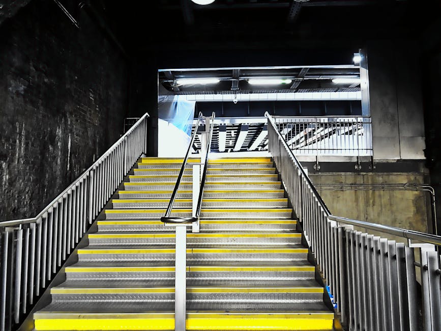A top-down view of a staircase within a building, showing a metal handrail running along the center of the stairwell, which has concrete steps with visible wear and light dust. The staircase is enclos