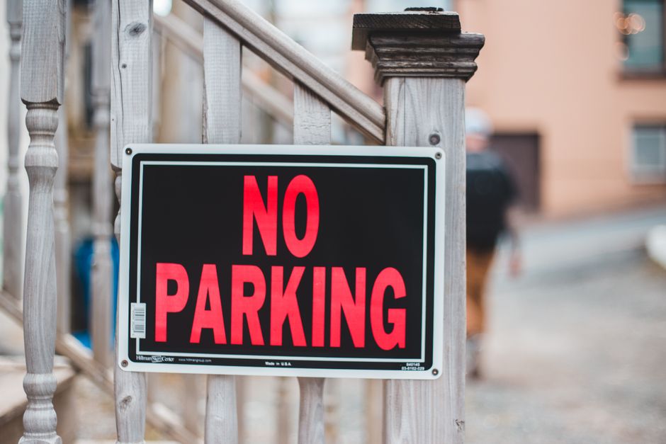 A close-up image of a rectangular black metal sign with gold lettering that reads 'NO PARKING' mounted on a light turquoise wooden wall composed of vertical planks. The sign is secured with two small 