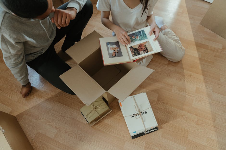 Two individuals are seated on a light wooden floor during a home relocation, with an open cardboard box between them containing framed photographs. One person, wearing a white top and beige pants, is 