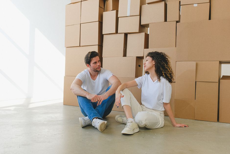 Two individuals are seated on a light wooden floor during a home relocation, with an open cardboard box between them containing framed photographs. One person, wearing a white top and beige pants, is 
